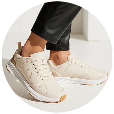 Beige Sneakers mit weißen Sohlen, getragen von Person in schwarzen Hosen, Nahaufnahme der Füße auf hellem Boden