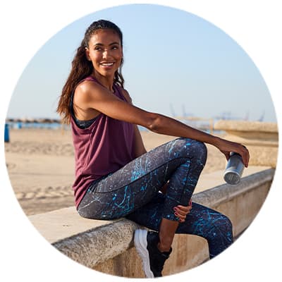 Frau in Sportkleidung sitzt auf einer Mauer am Strand und hält eine Wasserflasche.