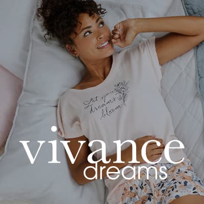 vivance dreams – Frau liegt entspannt auf dem Bett und trägt ein Schlafshirt mit dem Schriftzug „Let your dreams bloom“