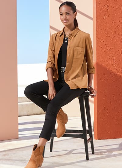 Personne assise sur un tabouret, chemise camel ouverte sur top noir, pantalon noir et bottines camel, fond terrasse ensoleillée