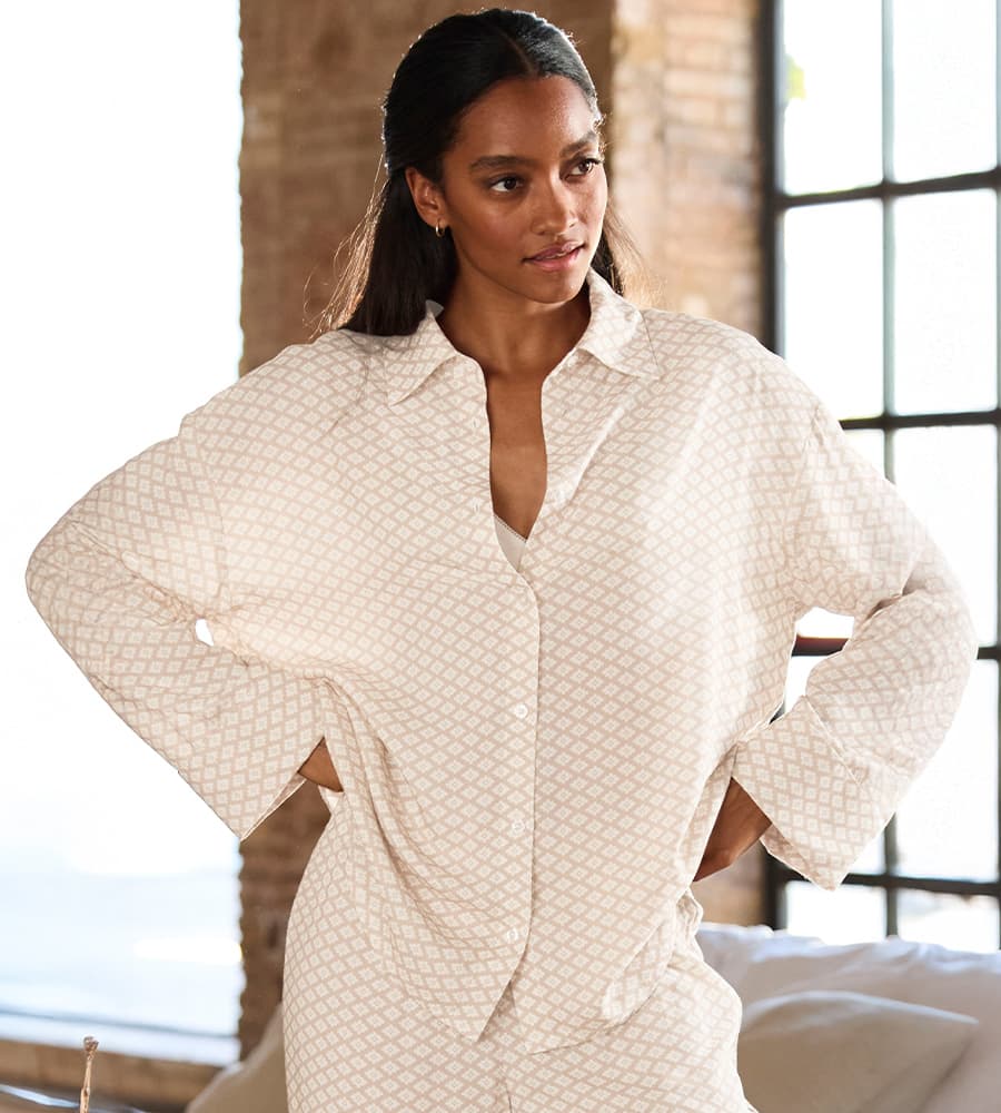 Nachtwäsche – Frau trägt beige gemustertes Nachtwäsche-Set, steht mit Händen in den Hüften vor einem Fenster.