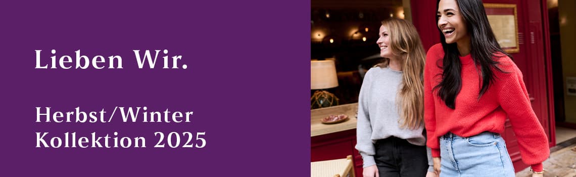 Lieben wir. Herbst/Winter Kollektion 2025 – Zwei Frauen in gemütlichen Pullovern, eine grau, eine rot, stehen nebeneinander