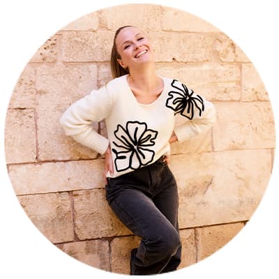 Femme en pull blanc avec motifs de fleurs noires, posant contre un mur de pierre