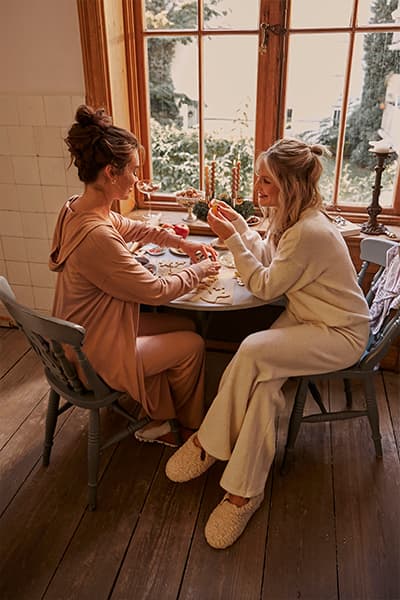 Zwei Frauen in gemütlichen Outfits sitzen am Tisch und spielen ein Brettspiel, durch das Fenster sieht man grüne Bäume.