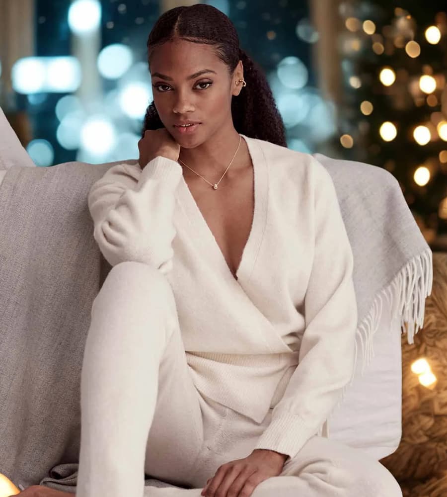 Frau in cremefarbenem Pullover und Hose sitzt auf einem Sofa, Weihnachtslichter im Hintergrund