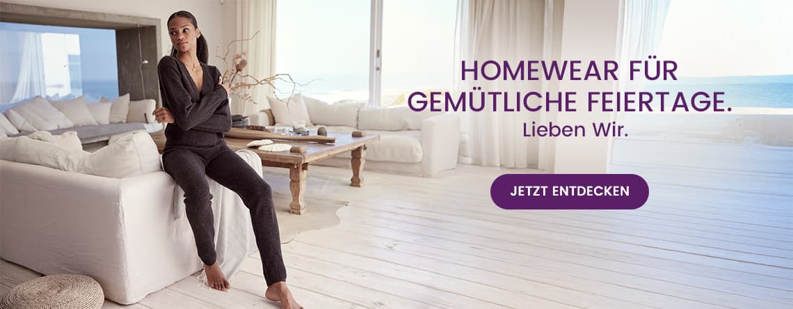 Homewear für gemütliche Feiertage. Lieben Wir. Jetzt entdecken – Frau in dunkelgrauem Hausanzug sitzt auf weißem Sofa, Meerblick im Hintergrund