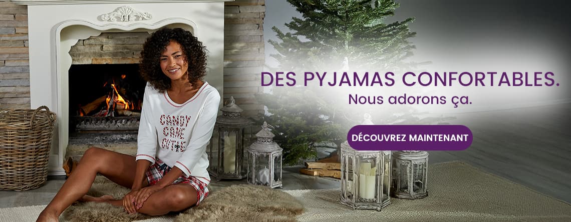 Des pyjamas confortables. Nous adorons ça. Découvrez maintenant – Femme assise en pyjama devant une cheminée, sapin de Noël à côté.