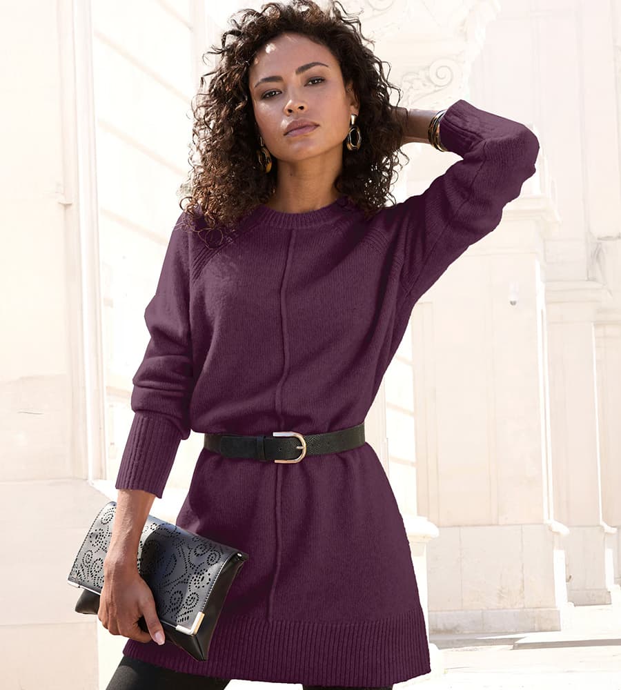 Frau in violettem Pulloverkleid mit Gürtel, schwarze Clutch in der Hand, steht vor einem hellen Gebäude.