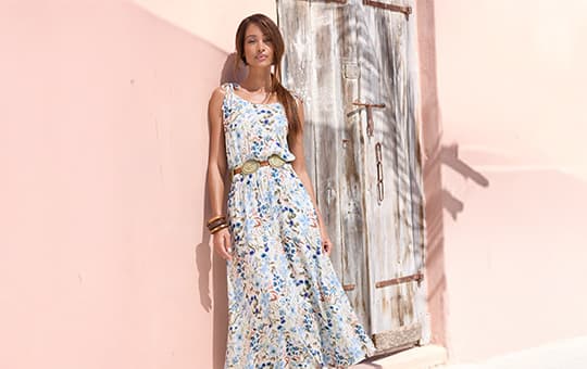 Frau in hellblauem Sommerkleid mit Blumenmuster lehnt an rosa Wand neben Holztür.