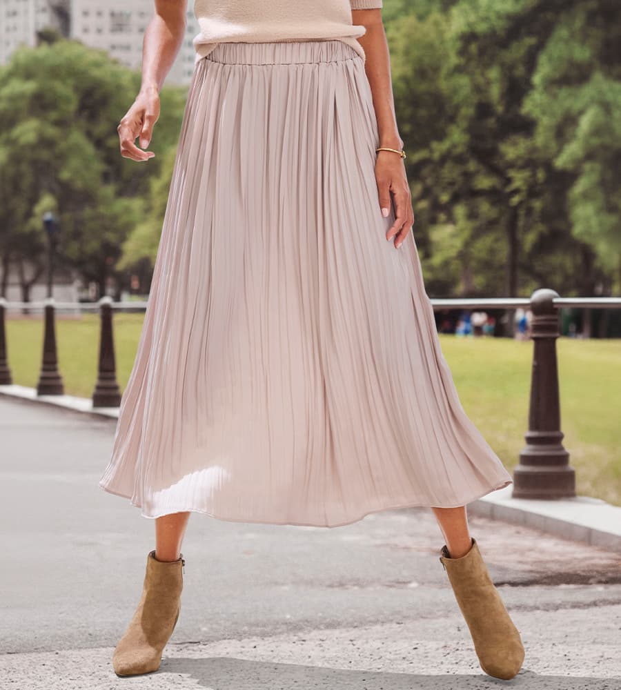 RÖCKE – Frau trägt einen fließenden, plissierten Rock und Stiefeletten, im Freien mit Bäumen im Hintergrund.