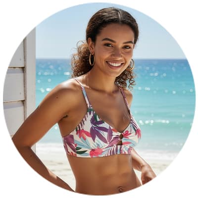 Femme en haut de bikini floral, posant devant une plage ensoleillée avec mer turquoise en arrière-plan.
