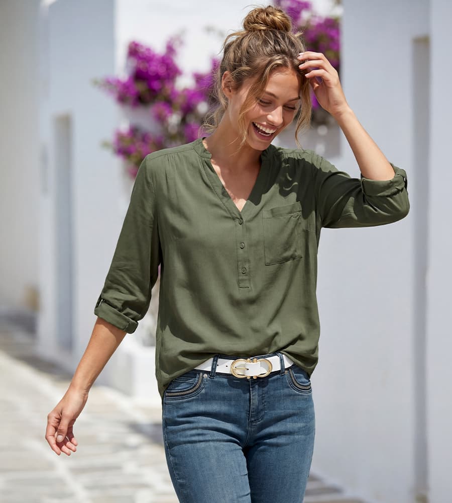 Frau in olivgrüner Langarmbluse und Jeans mit weißem Gürtel geht eine helle Gasse entlang