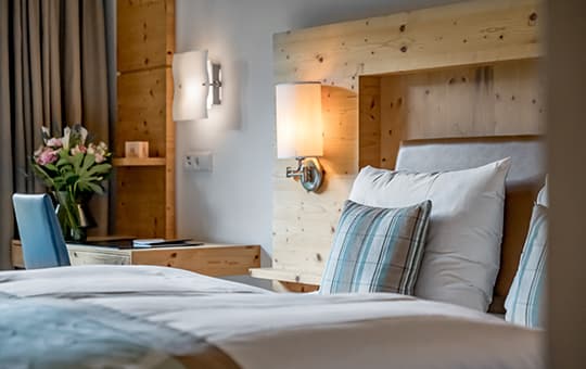 Doppelbett mit karierten Kissen, Holz-Kopfteil mit Wandleuchten, daneben Schreibtisch mit Stuhl und Vase mit Blumen