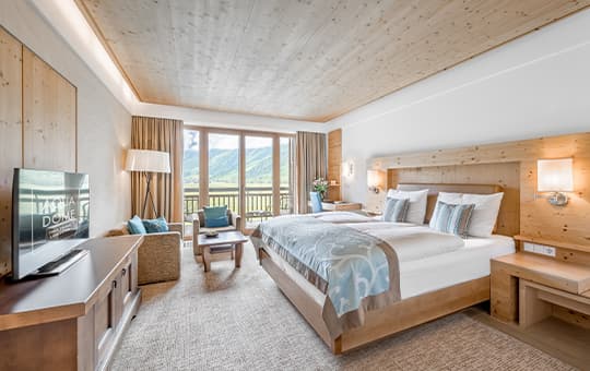 Helles Hotelzimmer mit Doppelbett, Sitzgruppe und Balkonblick auf grüne Berge durch große Fenster