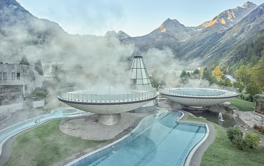 Dampfbad mit runden Außenbecken und langem Pool, umgeben von Bergen und grünen Hängen im Tal