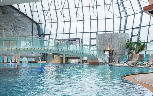 Großes Hallenbad mit Glasdach, geschwungener Brücke über dem Wasser und Liegestühlen am Beckenrand