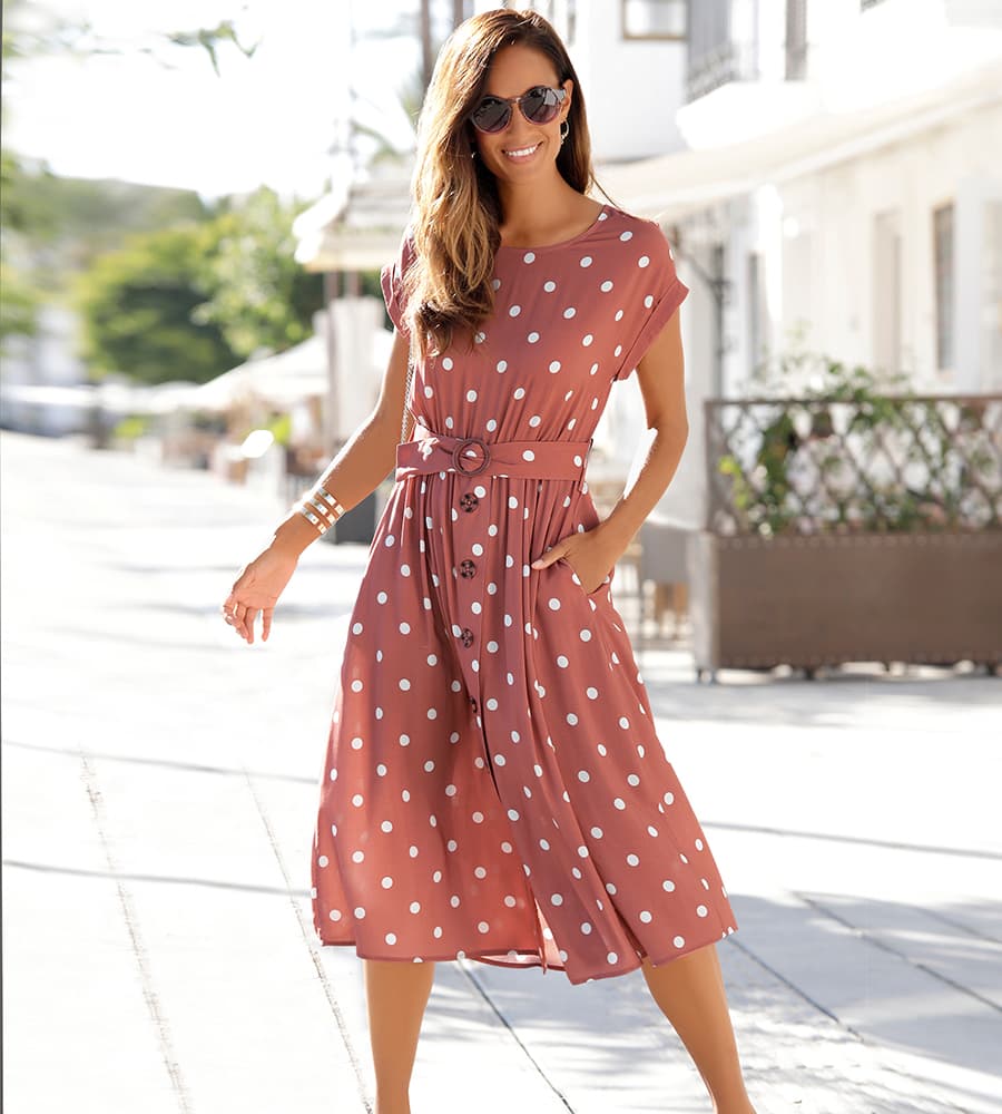Robe midi rose à pois blancs avec ceinture et boutons, portée par une femme marchant en extérieur