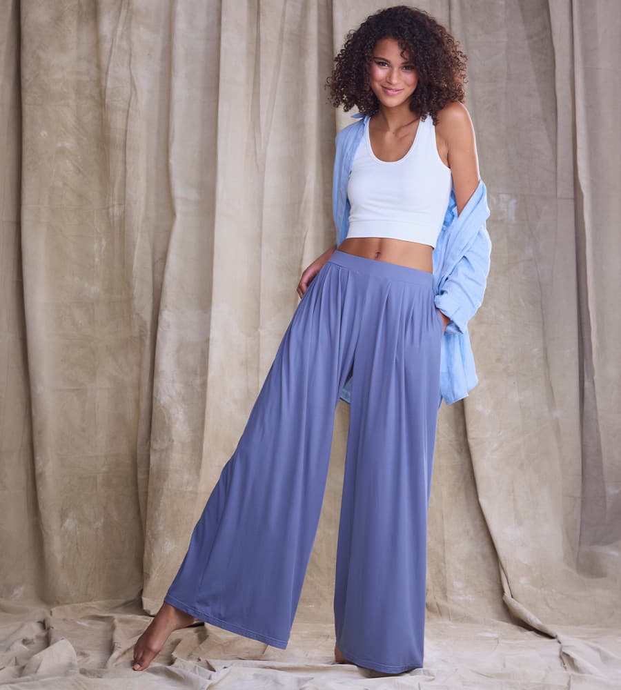 Femme debout en pantalon large bleu, débardeur blanc et chemise ouverte bleue, devant un rideau beige