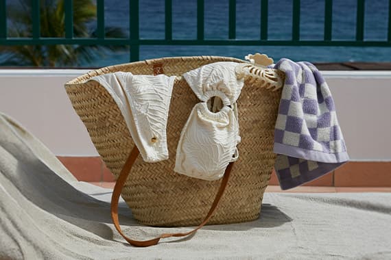 Geflochtene Strandtasche mit weißem Bikini und lila kariertem Handtuch auf Liege vor Meer und Palmen