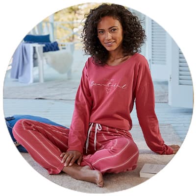 Frau in rosa Pyjama mit Schriftzug „Beautiful“ sitzt barfuß lächelnd auf einer Veranda