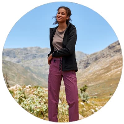Frau in Outdoorhose und Jacke steht entspannt in bergiger Landschaft und blickt in die Ferne