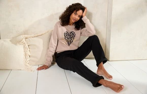 Frau sitzt barfuß auf hellem Boden, trägt Pullover mit Leoparden-Herz und dunkle Hose, lehnt an Kissen